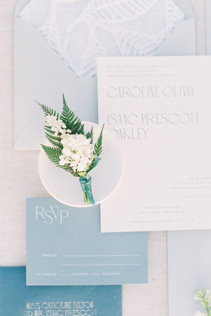 pale blue wedding invitations, white floral boutonniere on white dish, white florals with invites